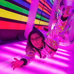 A surprised spacewoman floats in a room colored by a pink neon lighting effect. The left wall is painted in red, yellow, green and blue stripes. She is wearing glasses and reaching towards us.