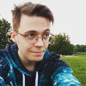 A man with a thoughtful expression smiles calmly, with a grey sky over trees and grass behind. He's wearing glasses and a hoodie in hues of blue with little white dots, recalling a night sky.