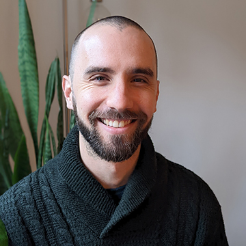 A man looks at us smiling widely and warmly, in an inside setting with a plant. He has a buzzcut haircut and a beard, and wears a high-collared woolen shirt.
