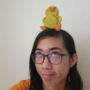 A headshot of a young man who, with pursed lips and a silly expression, looks up at a small plushie of a duck sitting on the top of his head.