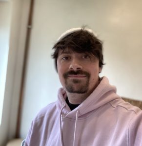 A man smiles, relaxed. He has unruly black hair and a goatee beard, and is dressed in a heavy pink hoodie and a pink bonnet. His picture was taken at home setting.