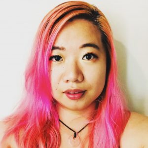 A woman with light pink hair down her shoulders looks at the camera. She wears a black tank top and a collar with a black string that holds a half transparent pink stone.