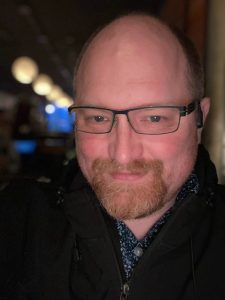 A man with a calm expression smiles warmly and happily, with dimples on his cheeks. He is bald and has a ginger circle beard. He's wearing a patterned shirt under a black jacket. The photo was taken at night. Behind him, we can glimpse a dark rooms with a few light spots.
